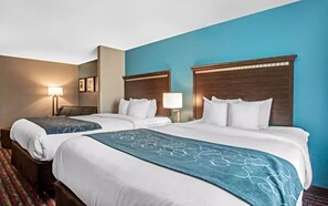 Habitación de hotel con dos camas, cada una con cabecero de madera, ropa de cama blanca y una manta azul. Entre las camas hay una mesita de noche con una lámpara, y un cuadro enmarcado en la pared.