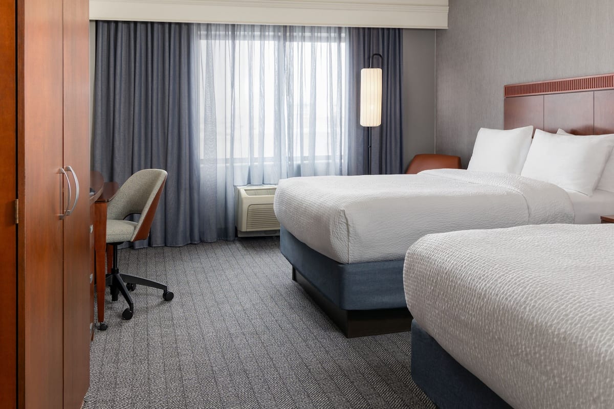 A hotel room with two beds, a desk, a chair, a lamp, and a window with curtains.