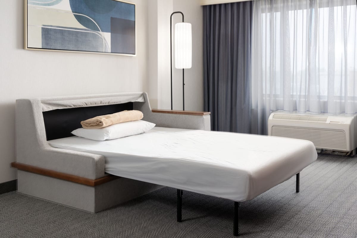A modern hotel room with a single bed, a bedside table, a floor lamp, a window with curtains, and a wall-mounted air conditioner.