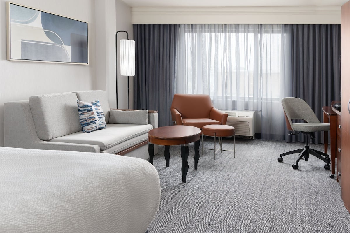 A hotel room with a sofa, two armchairs, a small table, a standing lamp, and a large window with curtains.