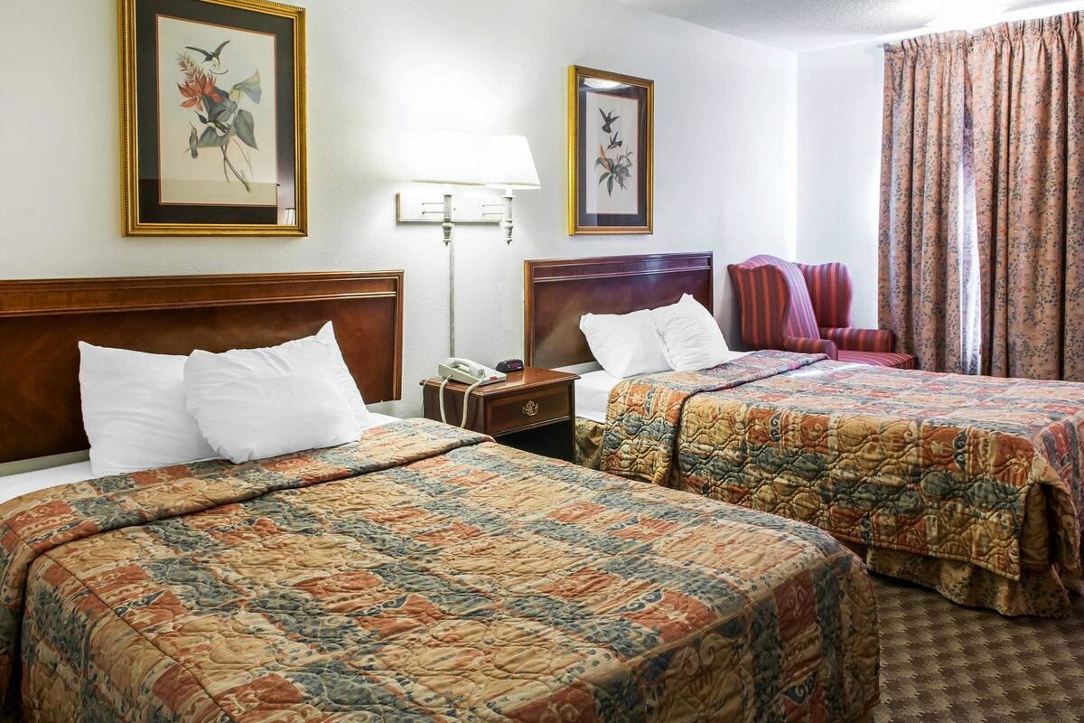 Quarto de hotel com duas camas, uma mesa de cabeceira, um abajur e cortinas florais.