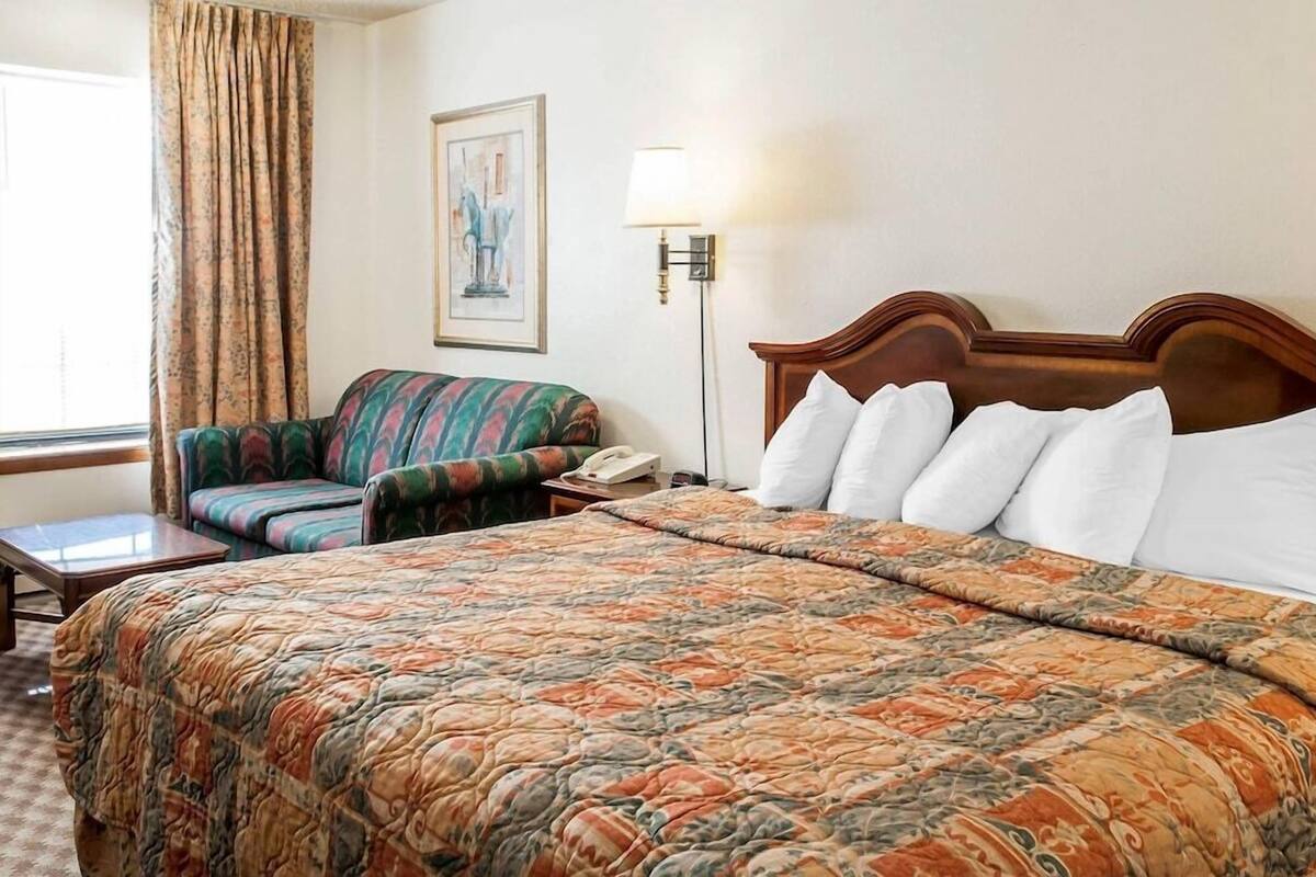 Quarto de hotel com cama, poltrona, escrivaninha, luminária e um quadro na parede.