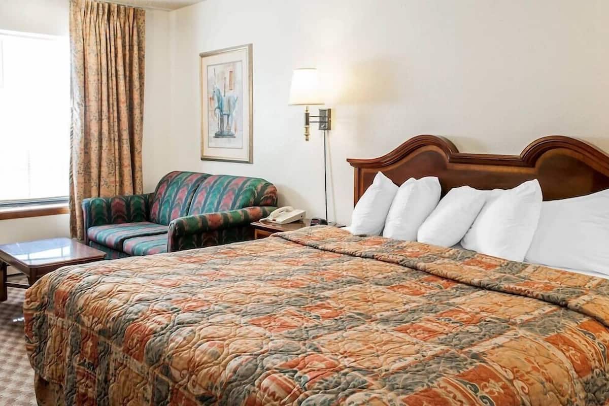 A hotel room with a bed, a chair, a desk, a lamp, and a painting on the wall.
