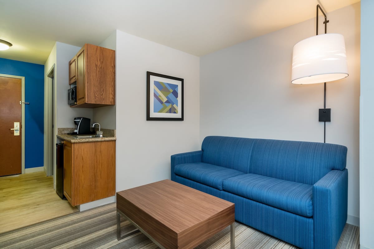 Una habitación moderna con un sofá azul, una mesa de centro de madera, una zona de cocina con microondas y una obra de arte abstracta enmarcada en la pared.
