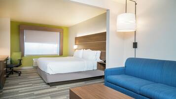 Quarto de hotel com cama, sofá azul, mesa de centro de madeira, escrivaninha com cadeira e janela com persianas.