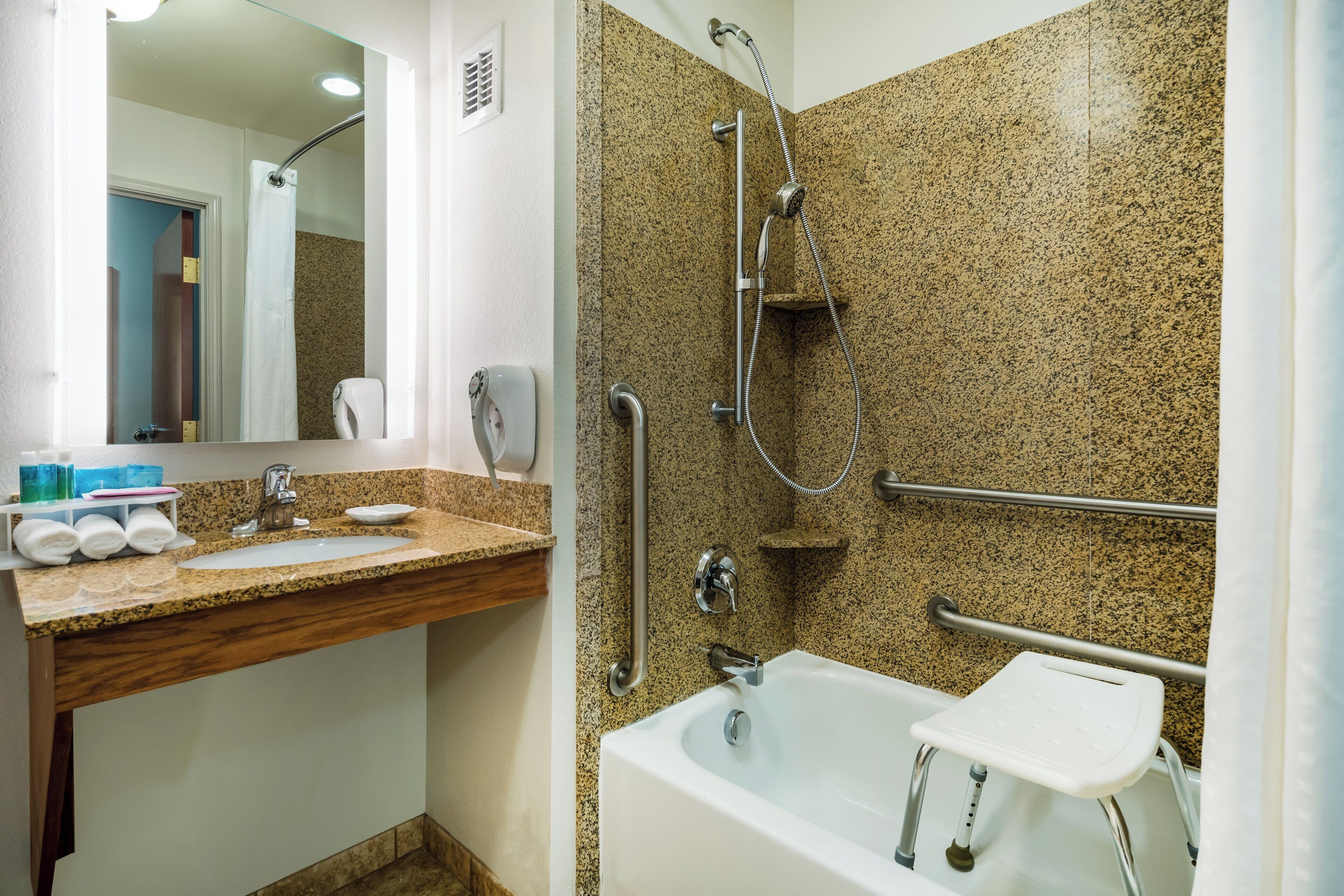 A bathroom with a granite countertop, a white sink, a shower with a handheld showerhead, and a grab bar.