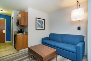 A compact living space with a kitchenette, a sofa, a coffee table, and a wall-mounted lamp. - Holiday Inn Express Hotel & Suites Rocky Mount by IHG (Rocky Mount)