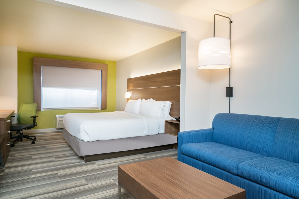 Habitación de hotel con cama, sofá azul, mesa de centro de madera, escritorio con silla y lámpara de pared.