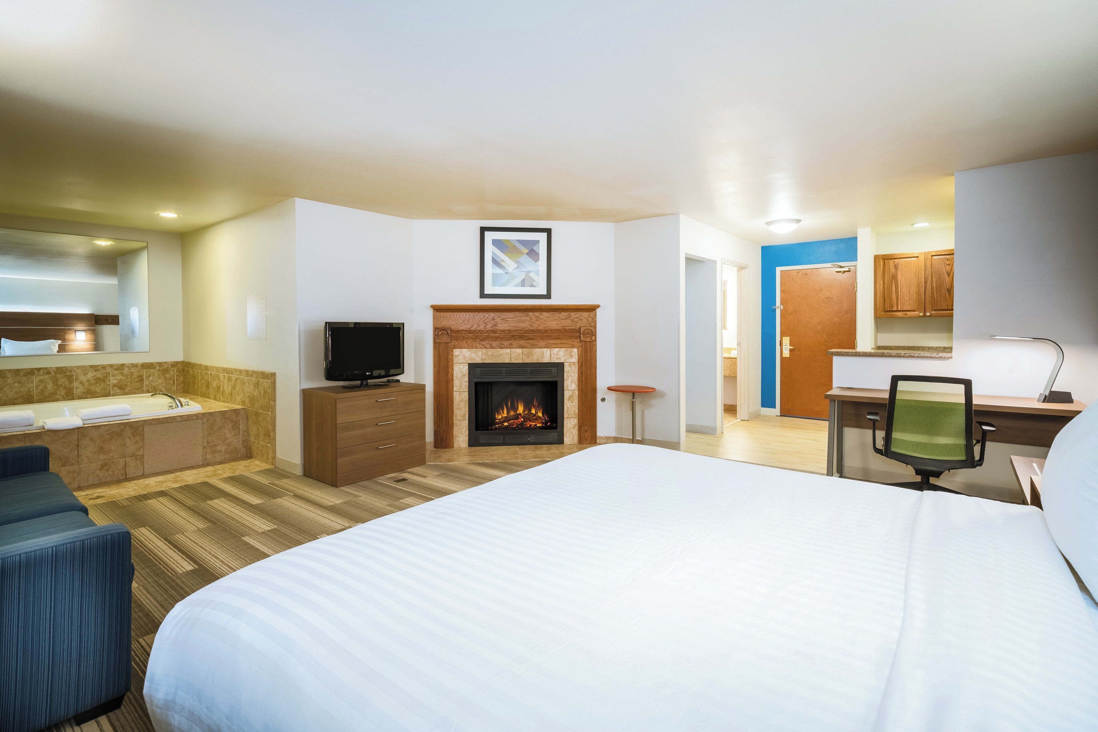A hotel room with a large bed, a fireplace, a television, a desk with a chair, and a bathroom with a bathtub and shower.