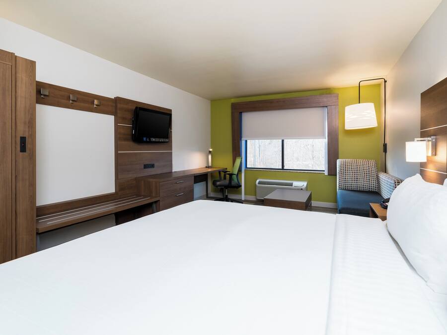 Quarto de hotel com uma cama grande, uma escrivaninha com cadeira, uma televisão fixada na parede e uma janela com persianas.