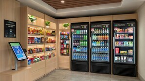 A self-checkout area with a digital screen, shelves stocked with snacks, and three beverage dispensers. - City Express By Marriott Rancho Cucamonga (Rancho Cucamonga)