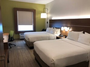 A hotel room with two beds, a desk, a chair, a window with blinds, and a wall-mounted lamp.