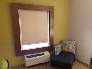 Een kamer met een groot raam voorzien van een beige zonnescherm, een witte airconditioning en een gestreept fauteuil, plus een klein tafeltje.