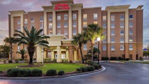 Un hotel Hampton Inn & Suites con entrada central, varios pisos y un césped bien cuidado con palmeras.