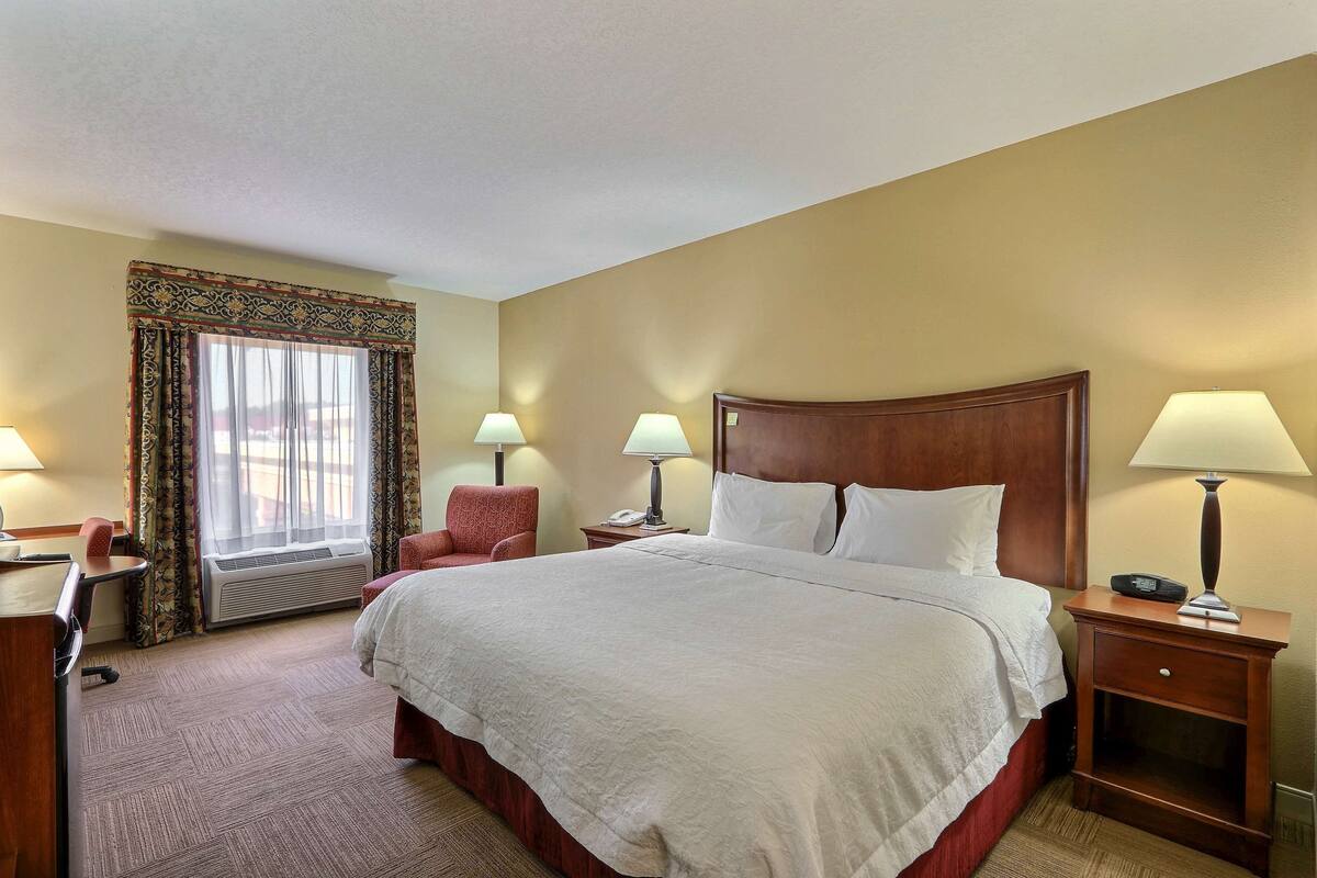 Habitación de hotel con una cama grande, dos mesitas de noche con lámparas, un escritorio con silla, una ventana con cortinas y un equipo de aire acondicionado.