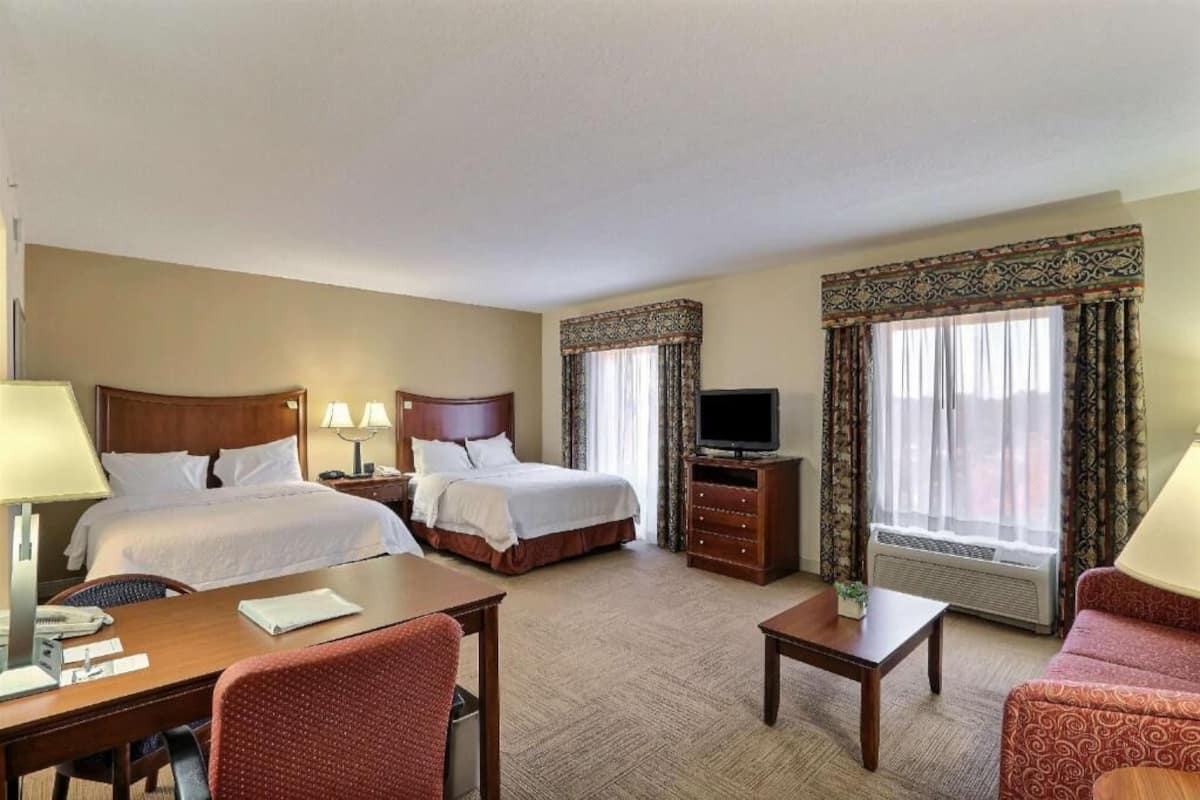 Habitación de hotel con dos camas, televisión, escritorio, silla, sofá y ventana con cortinas.