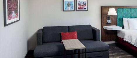 Quarto de hotel com sofá cinza escuro, mesa de cabeceira de madeira, cama com cabeceira branca e duas imagens emolduradas na parede.