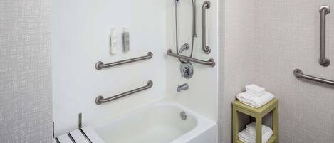 A bathroom with a white bathtub, grab bars, a small shelf with towels, and a shower with multiple nozzles.