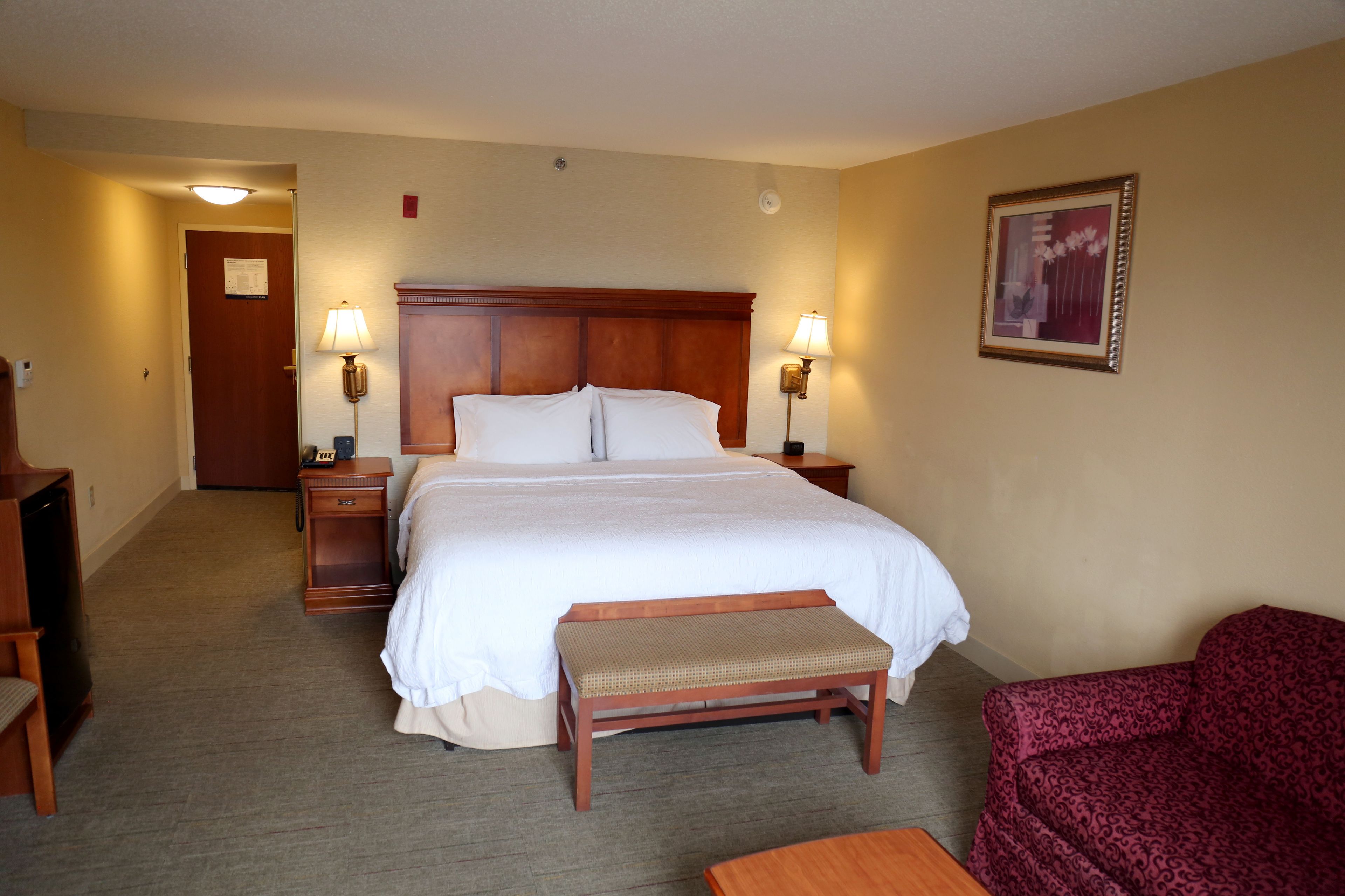 A hotel room with a large bed, two bedside tables with lamps, a bench, a chair, and a door leading to another room.
