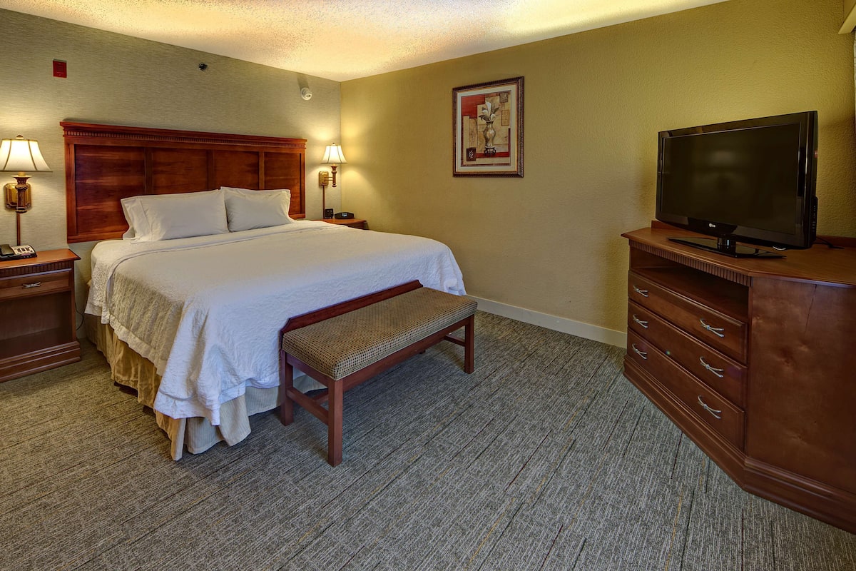 A hotel room with a large bed, a wooden bench, a television on a dresser, and a nightstand with a lamp.