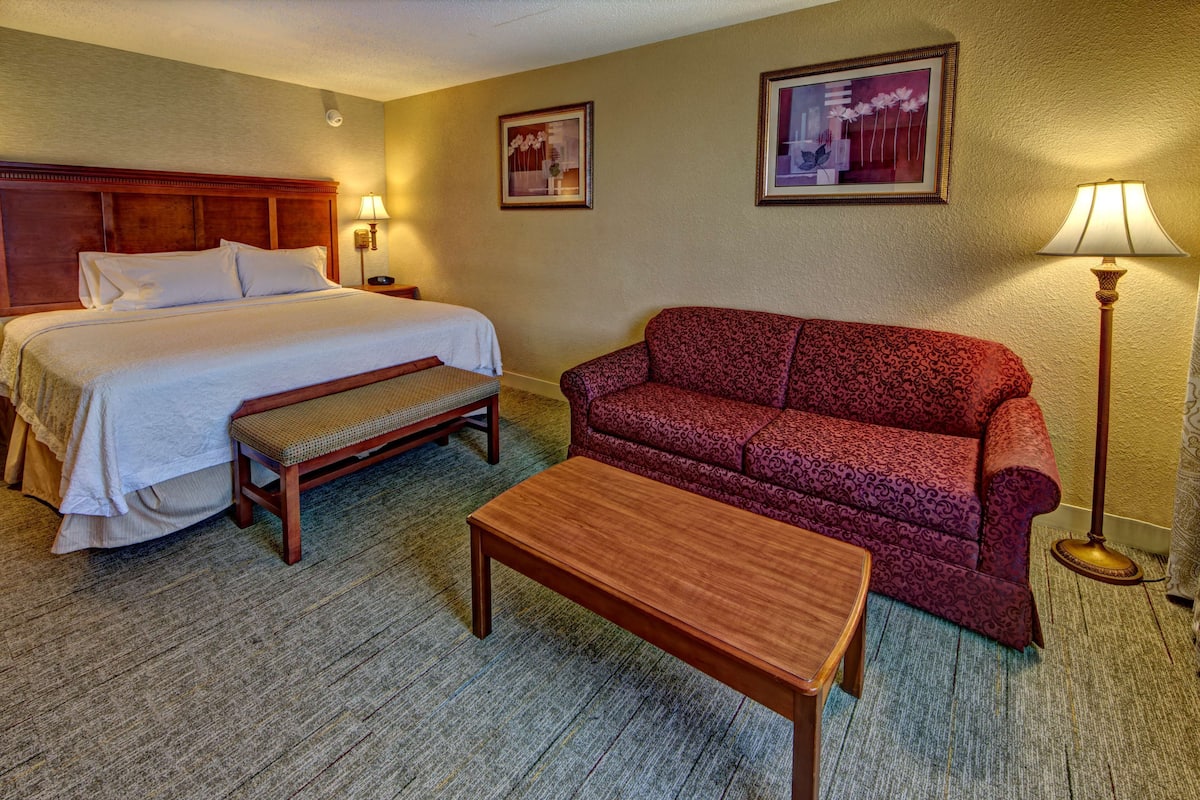 A hotel room with a bed, a red patterned sofa, a wooden bench, a small table, and two wall-mounted lamps.