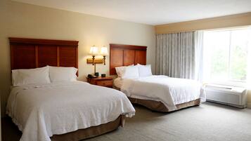 A hotel room with two beds, a nightstand with lamps, a window with curtains, and an air conditioning unit.