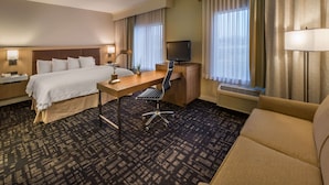A hotel room with a bed, desk, chair, TV, and large windows with curtains.