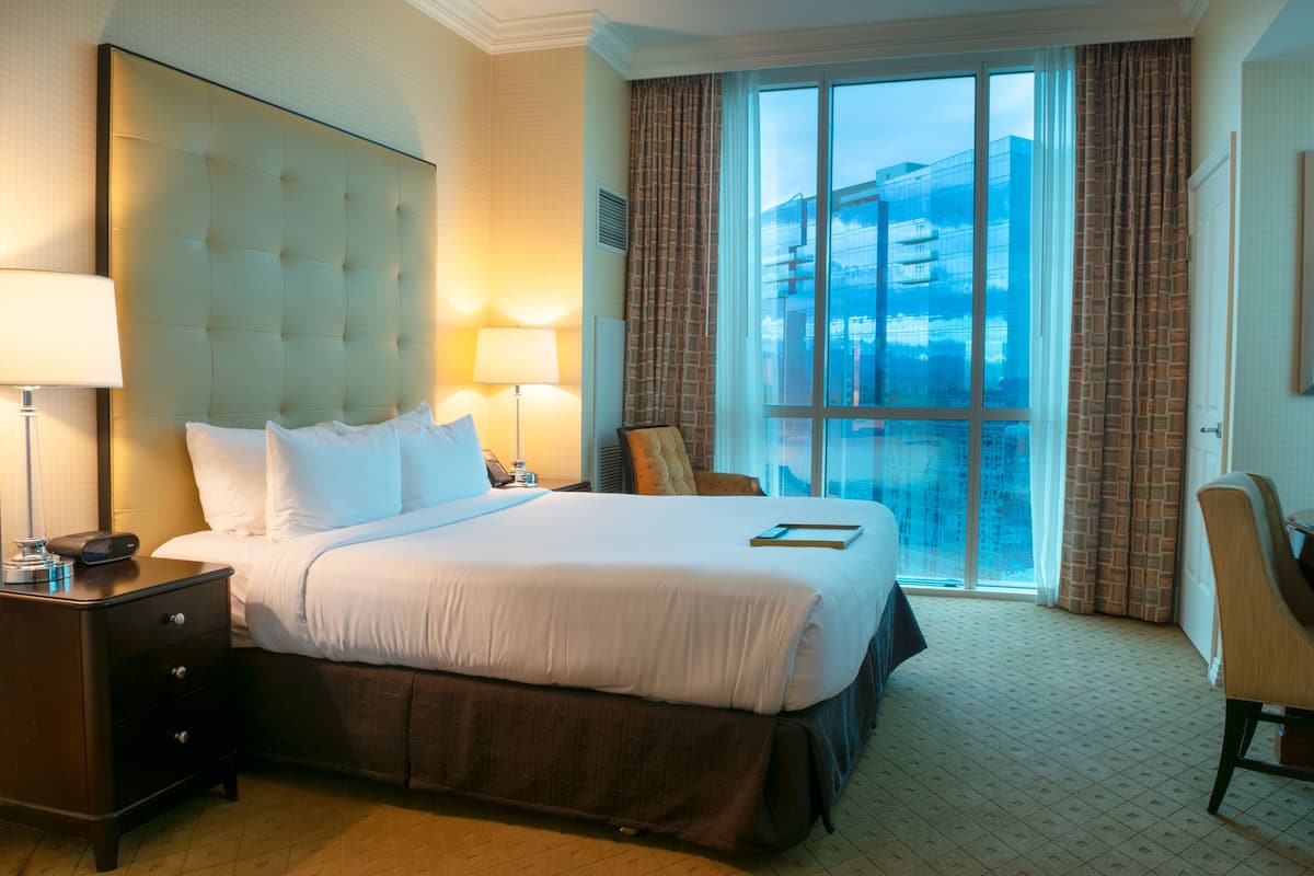 Habitación de hotel con una cama grande, dos lámparas de noche, una mesita, una silla y un ventanal con vistas a la ciudad.