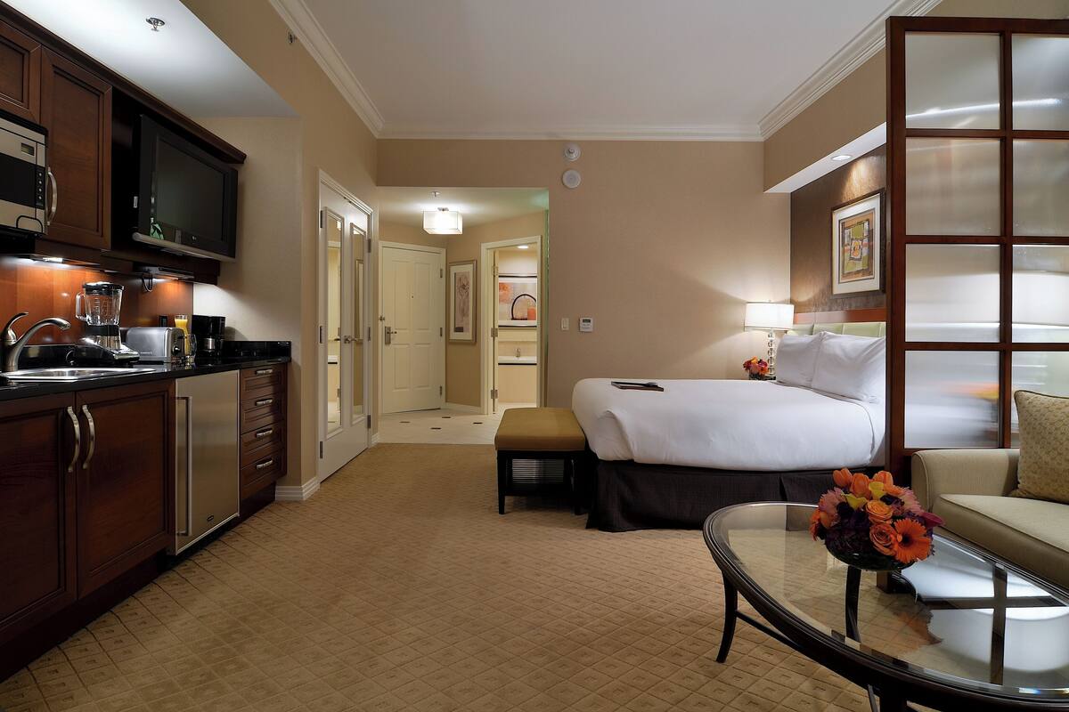Habitación de hotel con kitchenette, una cama, un sofá y una mesa de centro de vidrio con flores.