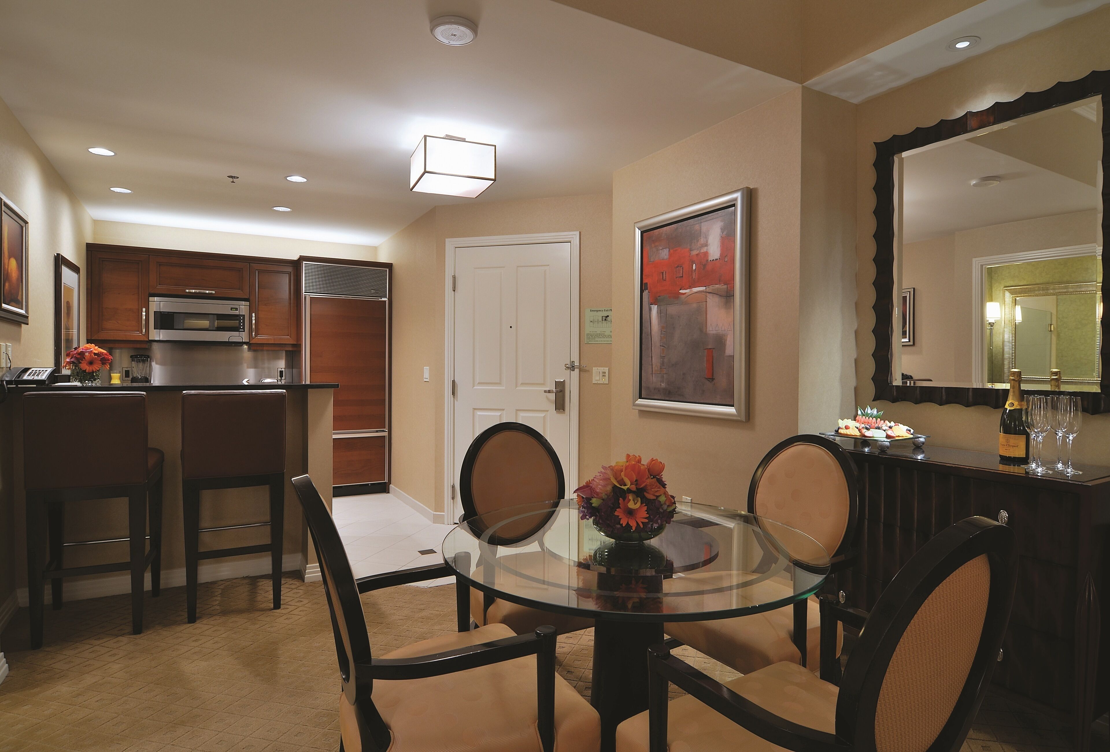 A dining area with a glass table, four chairs, a kitchen area with a microwave and refrigerator, and a large mirror.