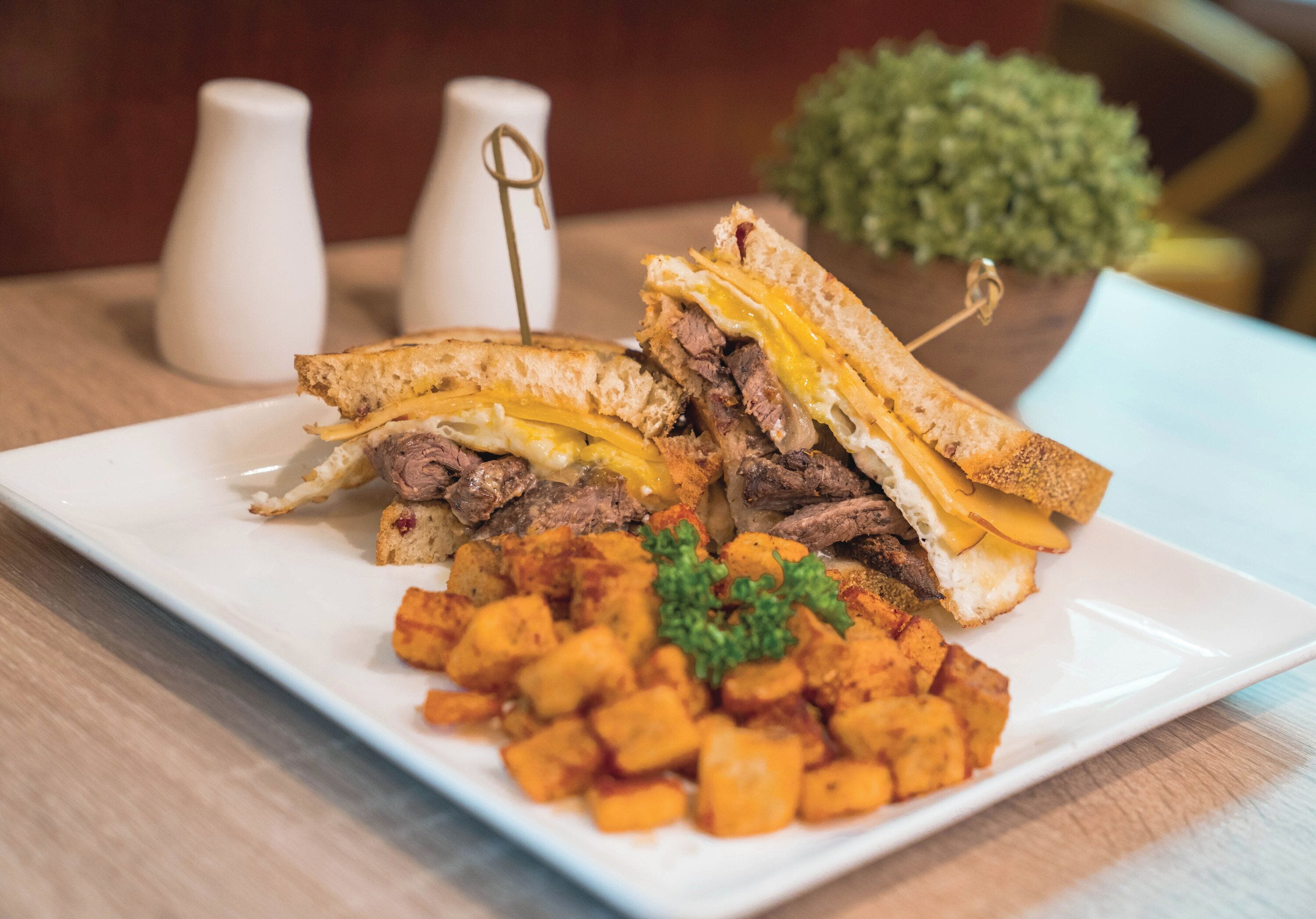 A plate with a sandwich cut in half, a side of roasted potatoes, and a small sprig of parsley.