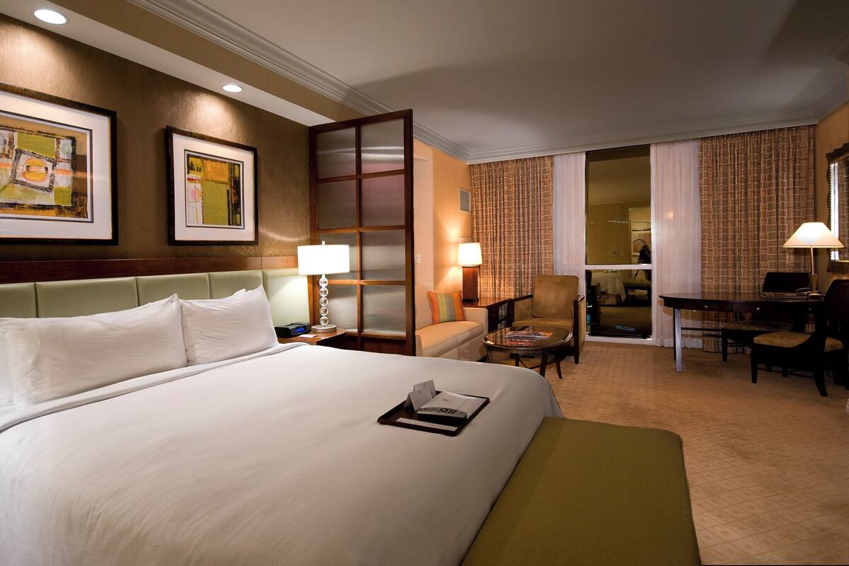 Habitación de hotel con una cama grande, un escritorio, una silla y un sofá. Hay dos lámparas en el escritorio y una ventana con cortinas.