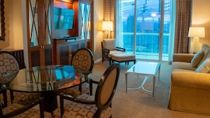 A hotel room with a glass dining table, cushioned chairs, a sofa, a TV, and a balcony with a city view.