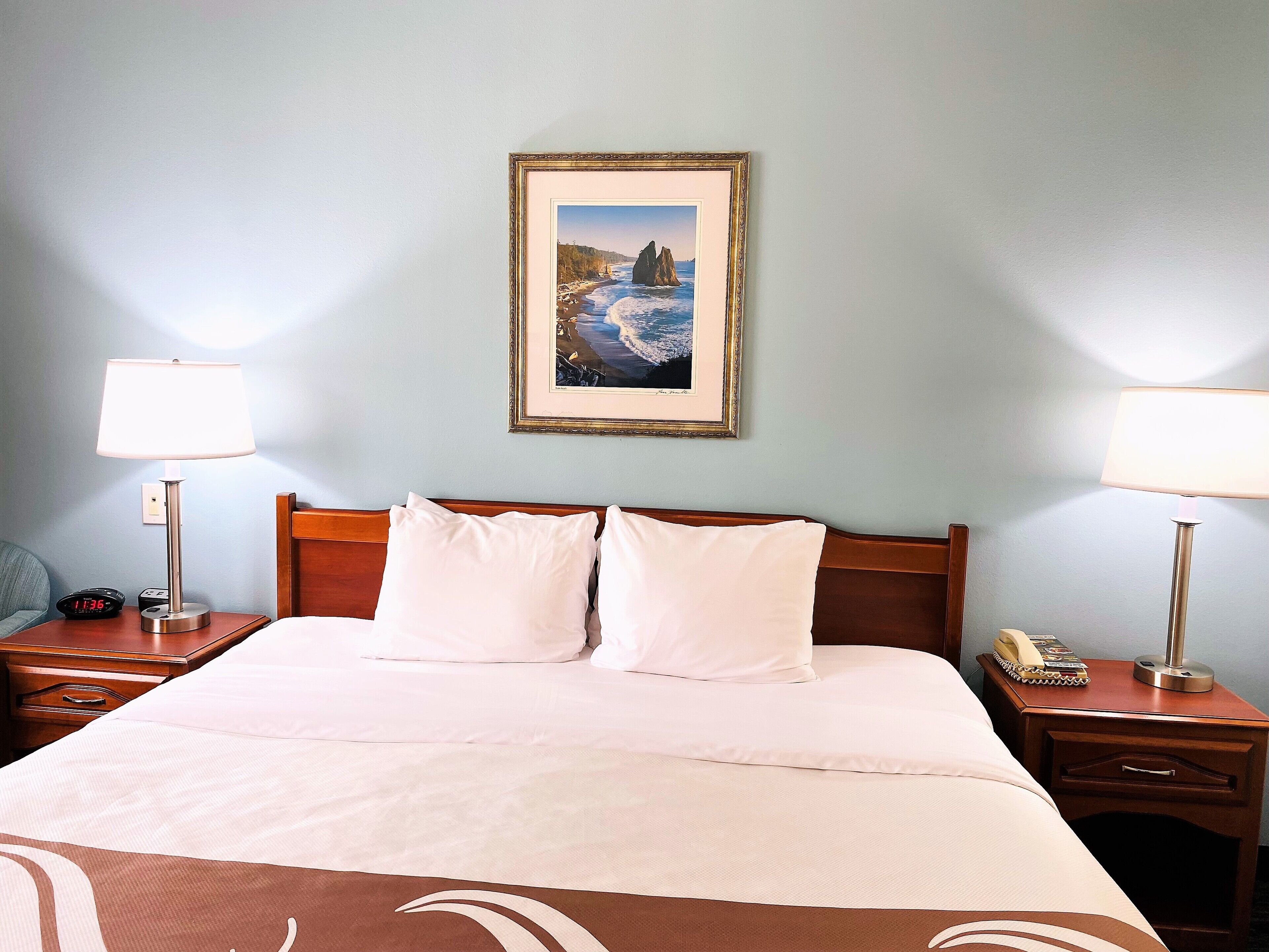 A hotel room with a bed, two bedside tables, a nightstand, a framed picture on the wall, and a clock on the nightstand.