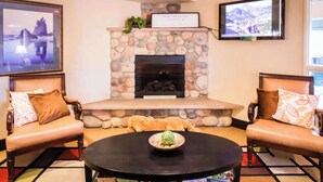 A cozy living room with a stone fireplace, two leather sofas with patterned cushions, a round coffee table with board games, and a television mounted on the wall. - Quality Inn & Suites Sequim at Olympic National Park (Sequim)
