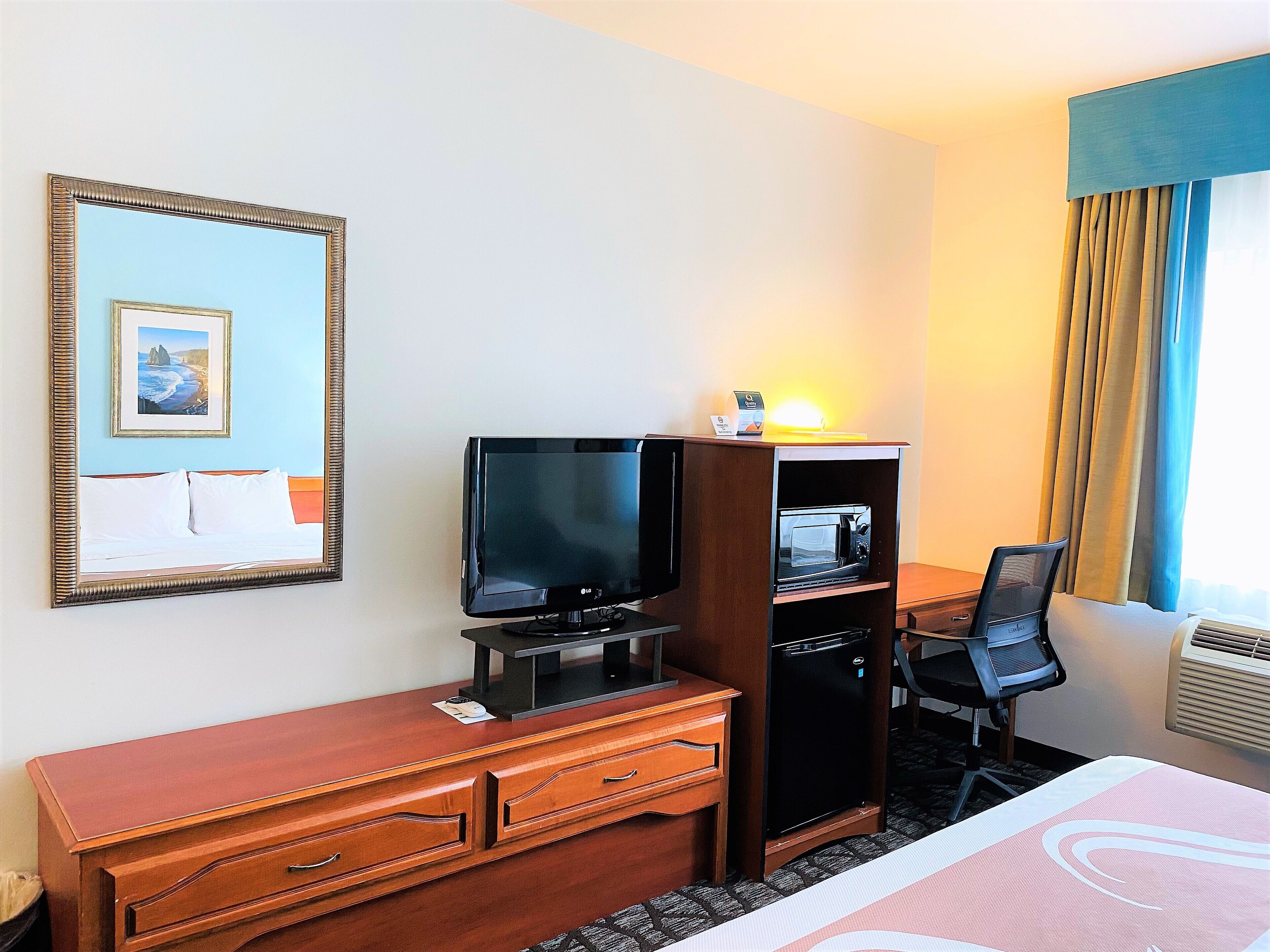 Una habitación de hotel con una cama, un escritorio con computadora, televisor, microondas, una silla y un aparador.