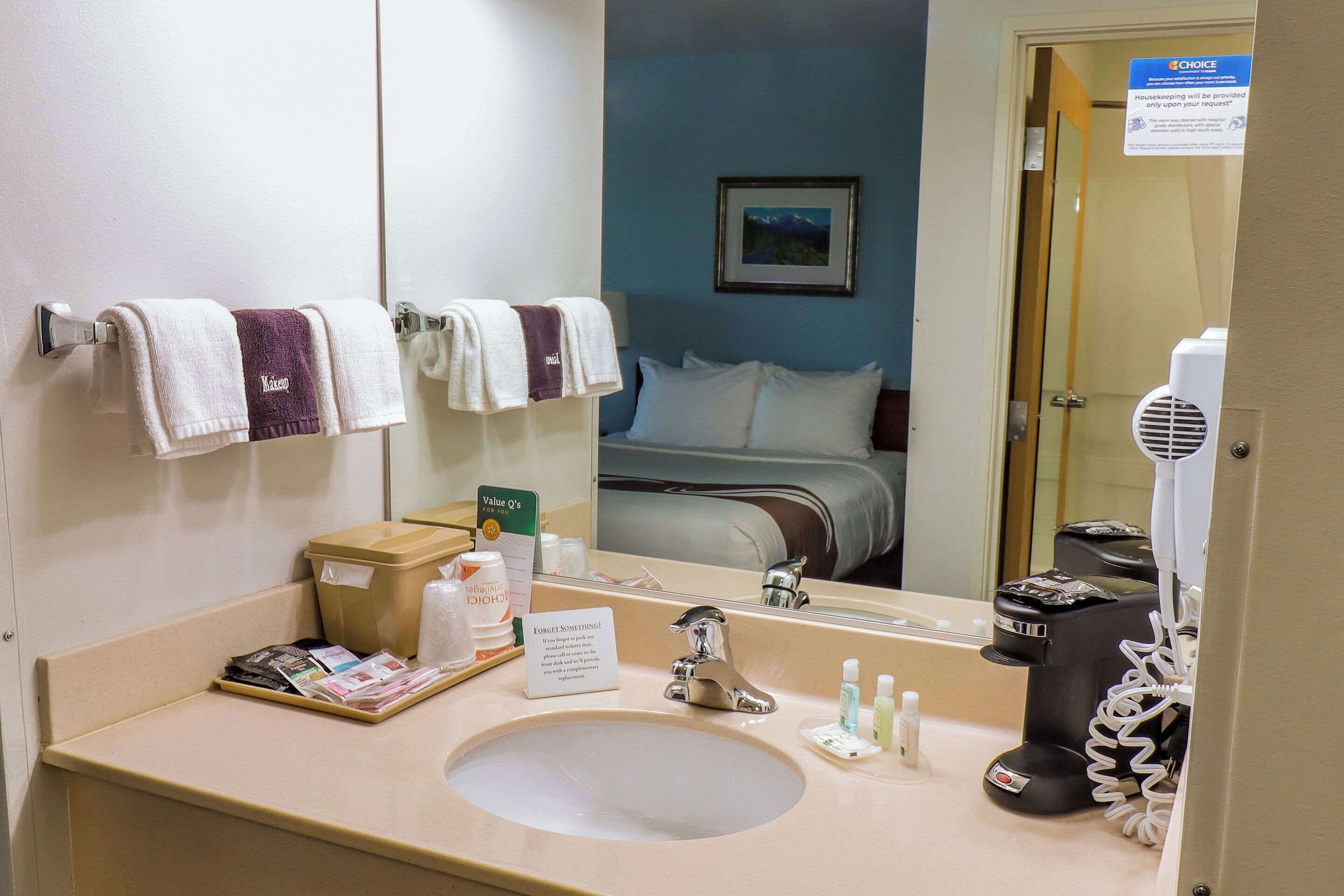 A hotel bathroom with a sink, mirror, towels, toiletries, and a bed visible through the door.