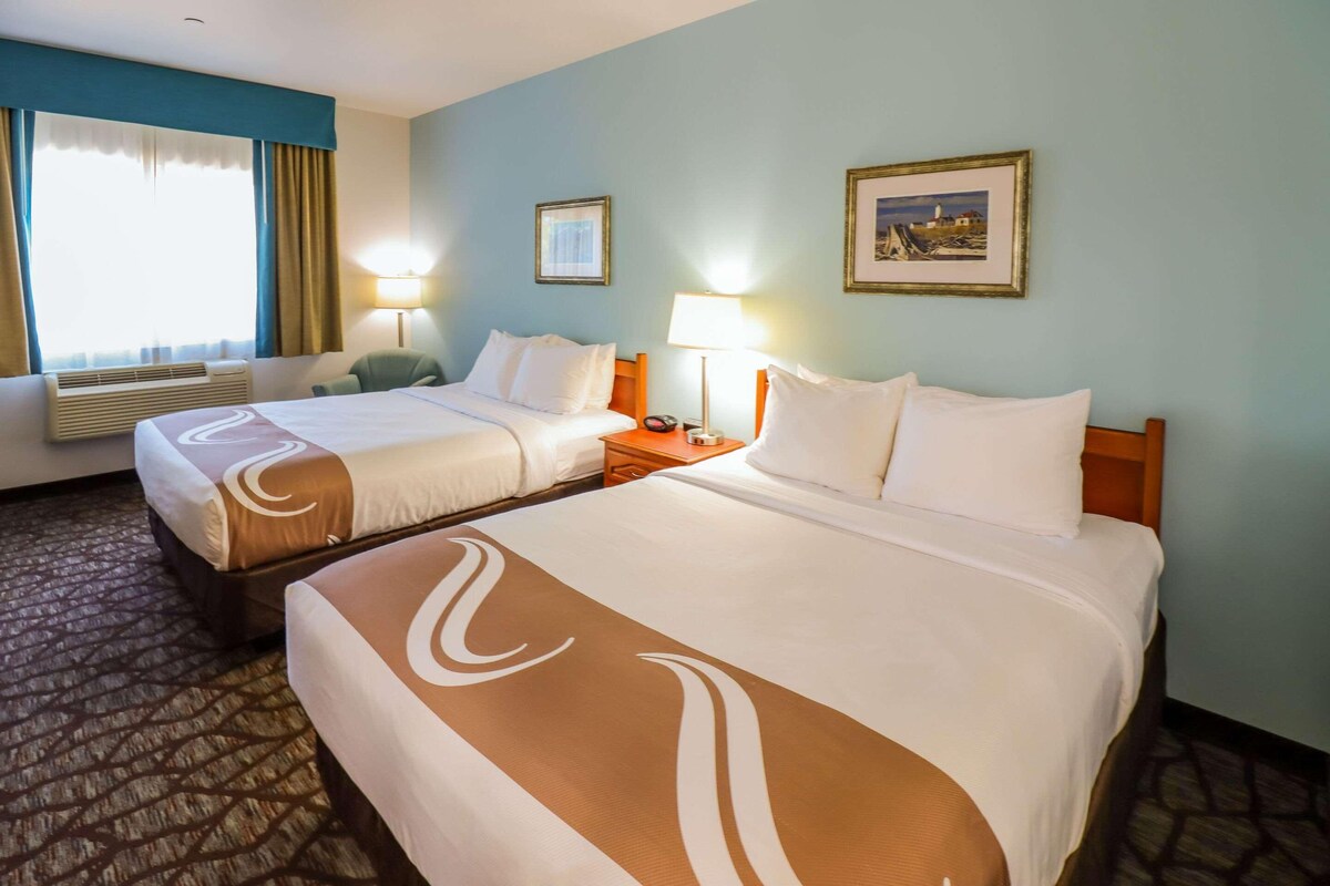 A hotel room with two beds, a window with curtains, a wall-mounted picture, and a nightstand with a lamp.