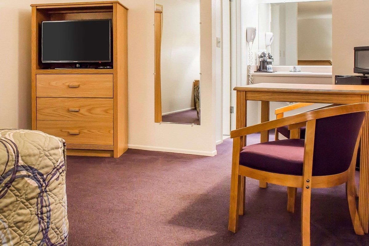 A room with a wooden TV stand, a mirror, a dining table with chairs, and a bed with a patterned blanket.