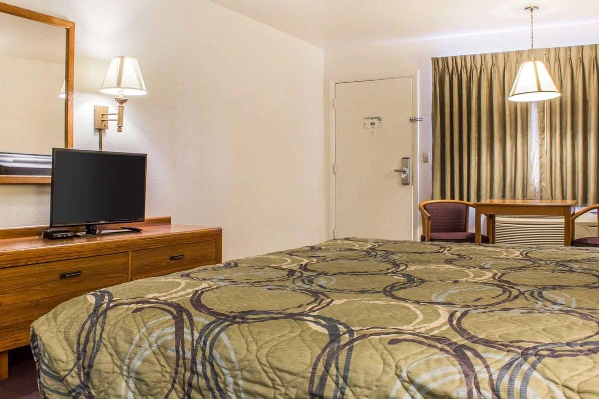 A hotel room with a bed, a desk with a television, a chair, a lamp, and a door.