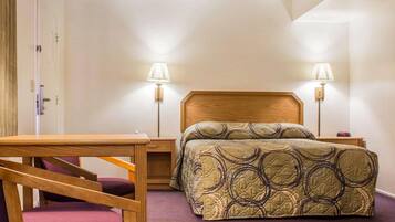 Una habitación de hotel con una cama, dos lámparas de noche, una mesita de noche de madera, una silla y una puerta.