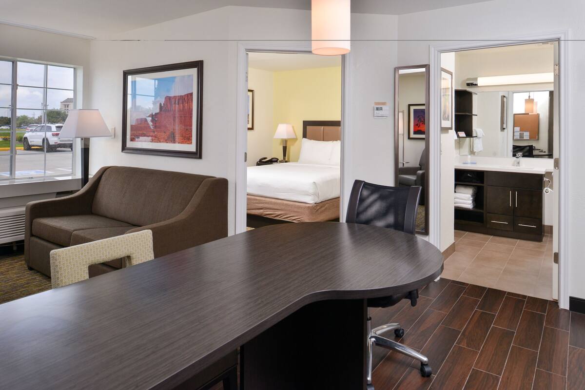 Ein Hotelzimmer mit einem großen Bett, einem Schreibtisch mit Stuhl, einer Sitzecke und einem Badezimmer mit Spiegel. Ein Fenster bietet einen Ausblick nach draußen.