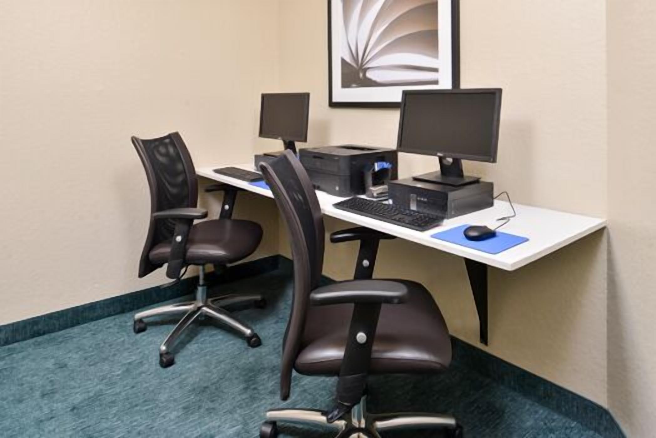 A workspace with two black office chairs, a white desk, two monitors, a keyboard, a mouse, and a framed picture on the wall.