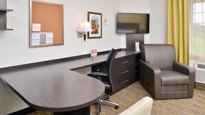 A hotel room with a desk, a comfortable armchair, a flat-screen TV mounted on the wall, a corkboard with notices, and a window with yellow curtains. - Candlewood Suites Beaumont by IHG (Beaumont)