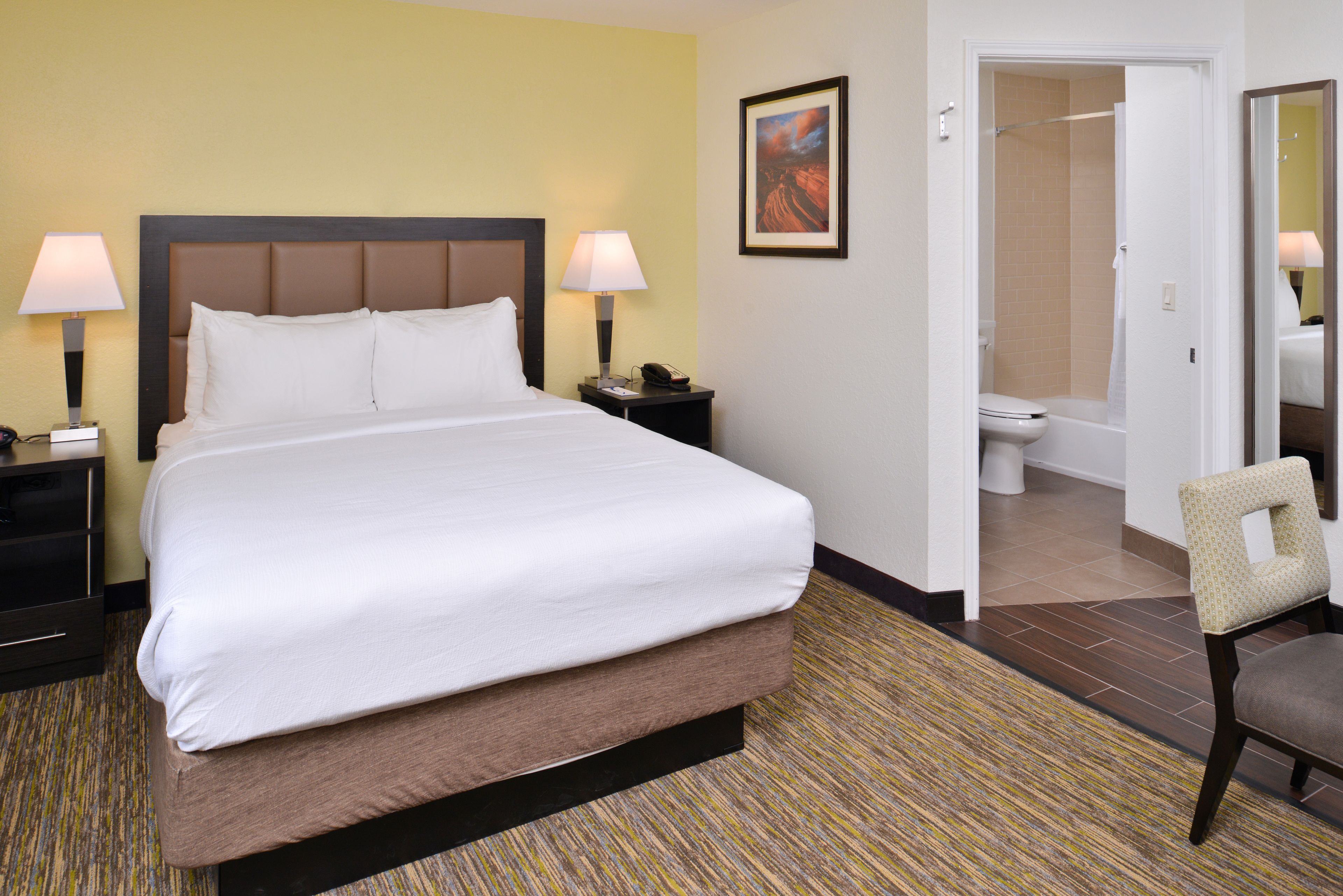 A hotel room with a large bed, two bedside lamps, a nightstand, a chair, a bathroom with a shower, and a framed picture on the wall.