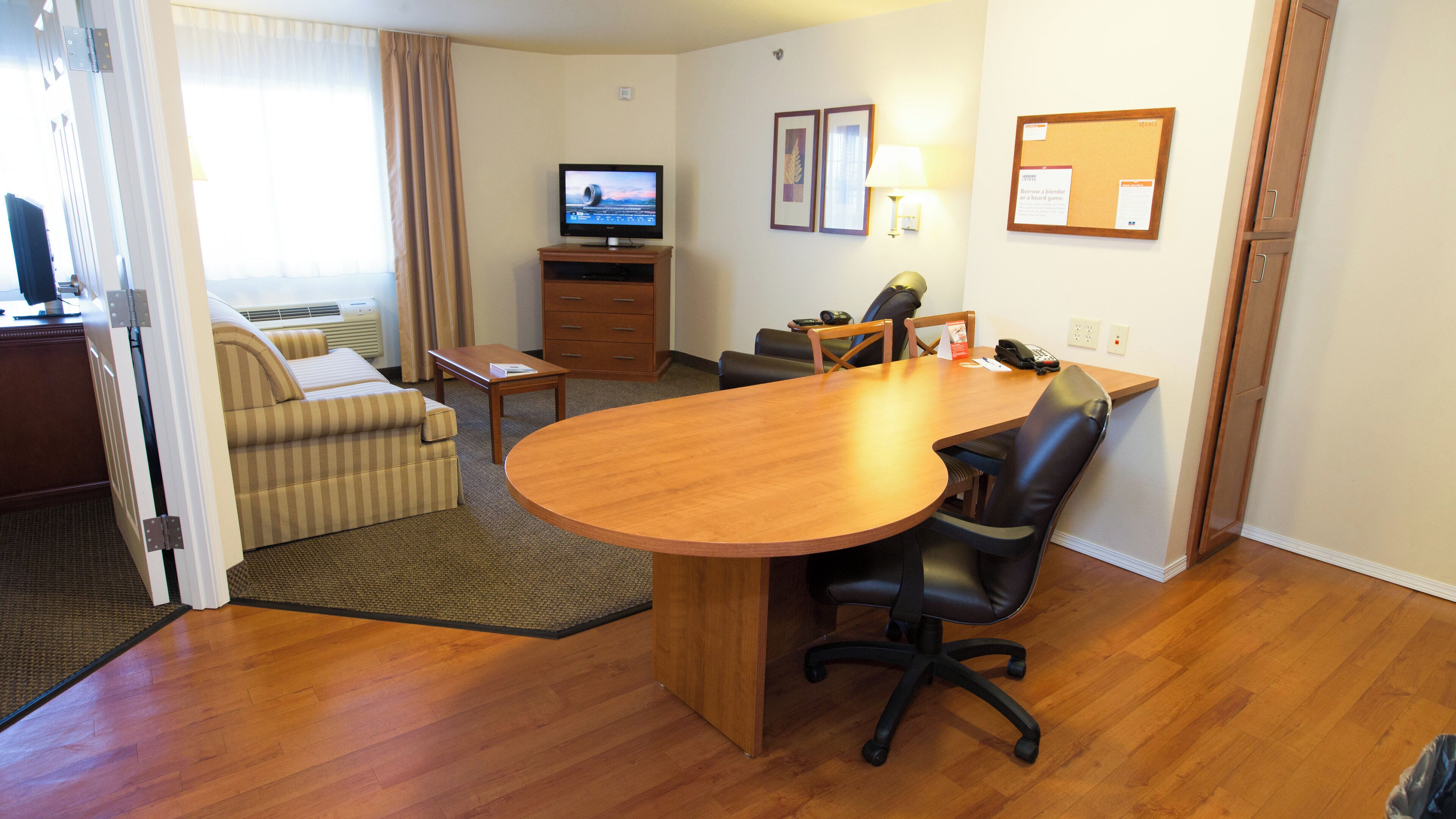 A hotel room with a wooden dining table, a sofa, a TV, and a desk with a lamp.