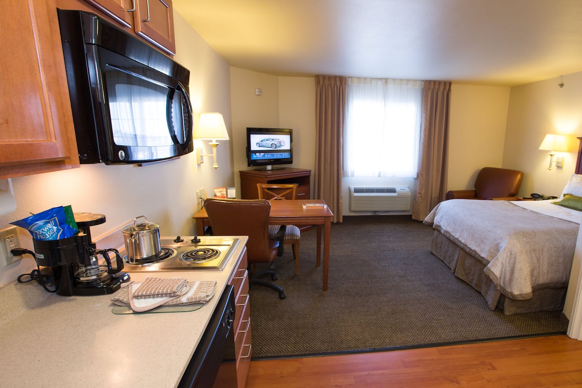 Ein Hotelzimmer mit Küchenzeile, einem Bett, einem Schreibtisch, einem Fernseher und einem fenster mit Vorhängen.