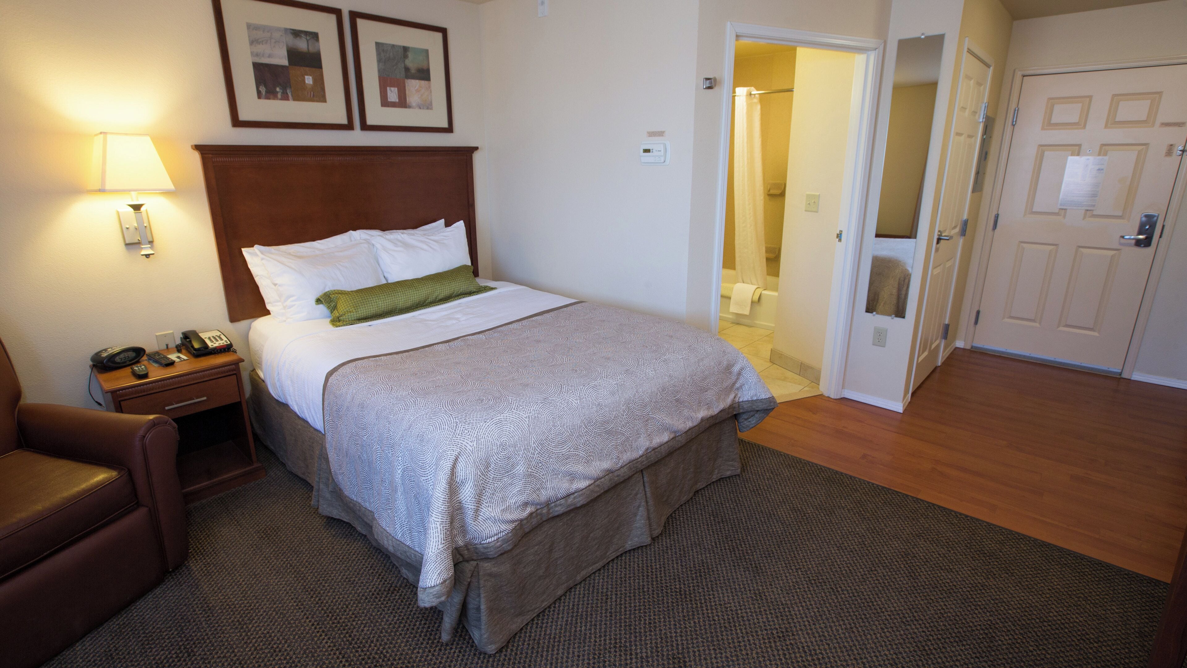A hotel room with a bed, a nightstand with a lamp, a chair, and a door leading to a bathroom.