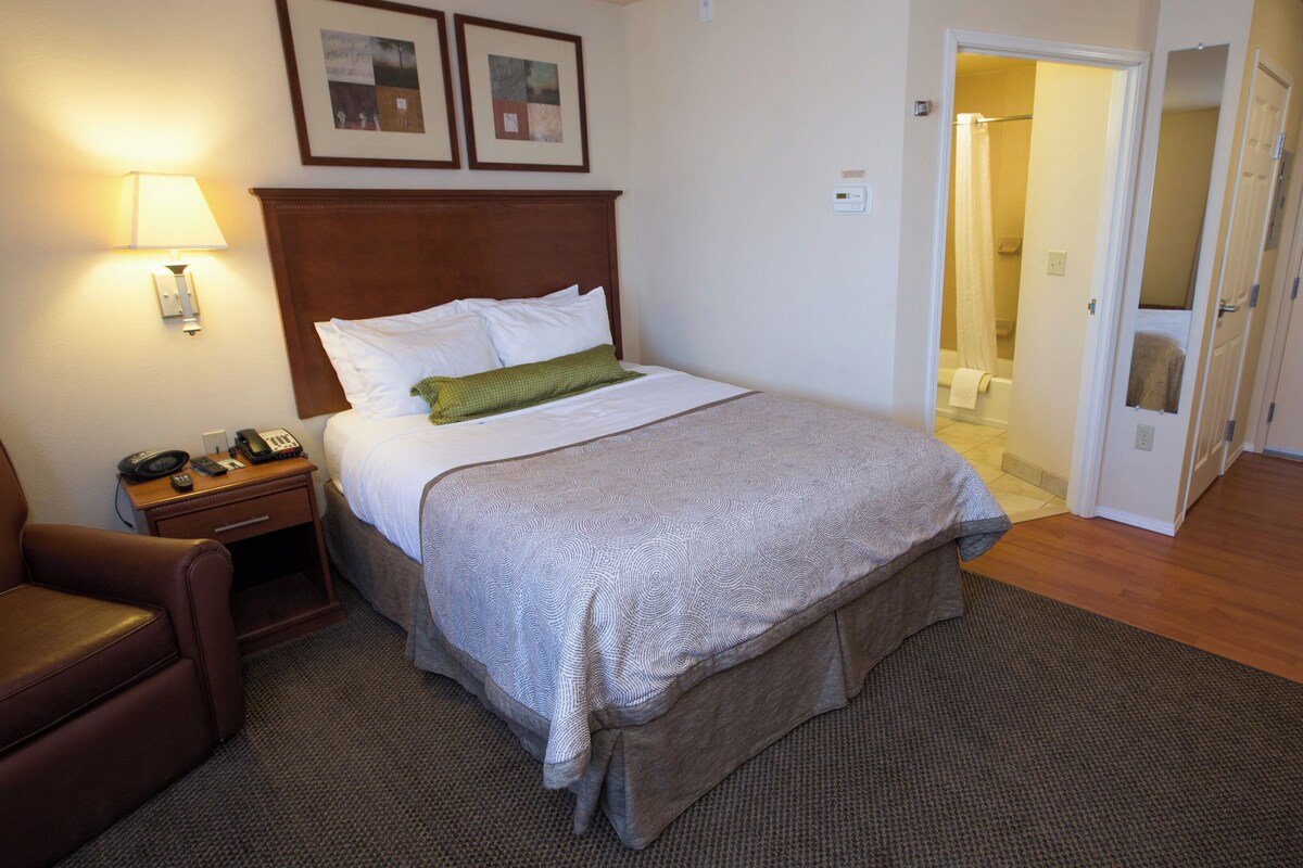 Une chambre d’hôtel comprenant un lit, une table de chevet avec une lampe, une chaise et une porte donnant sur une salle de bain.