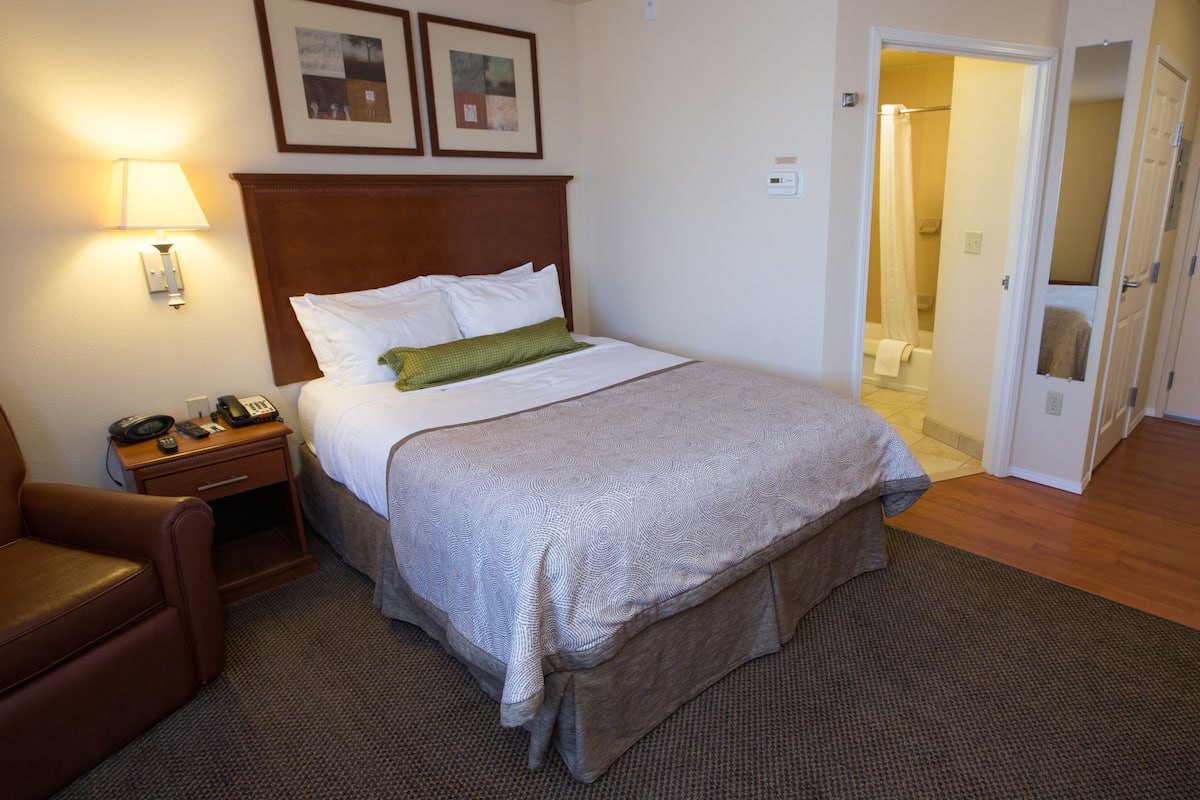 Ein Hotelzimmer mit einem Bett, einem Nachttisch mit Lampe, einem Sessel und einer Tür zu einem Badezimmer.